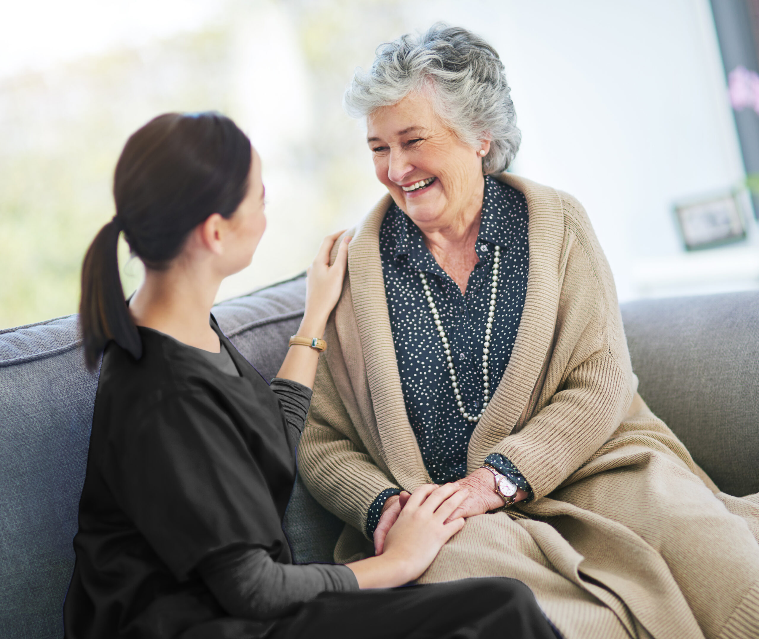 A compassionate caregiver and an older adult smile while talking together in a comfortable living room at home.
